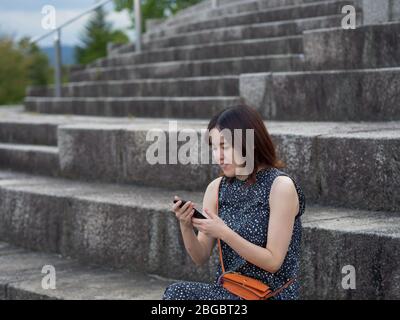 Eine Japanerin, die auf ihr Telefon schaute, während sie auf einem Steinschritt saß Stockfoto