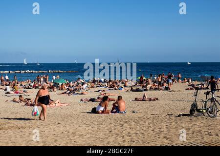 Blick auf viele Menschen, die sich auf Sand legen, sich sonnen und alte Frau am berühmten Strand 'La Barceloneta' in Barcelona spazieren gehen. Es ist ein sonniger Sumpfbecken Stockfoto