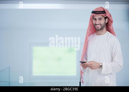 Junge moderne muslimische arabische Geschäftsmann in traditioneller Kleidung, während Smartphone zu Hause Stockfoto
