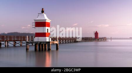 Lange Aufnahme des Roten Leuchtturms von Lignano Sabbiadoro bei einem kalten winterlichen Sonnenaufgang. Ruhiges Wasser unter einer Holzpromenade Stockfoto