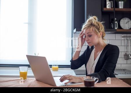 Junge Frau mit Kopfschmerzen während der Arbeit am Laptop. Sie sitzt an ihrem Küchentisch zu Hause. Stockfoto