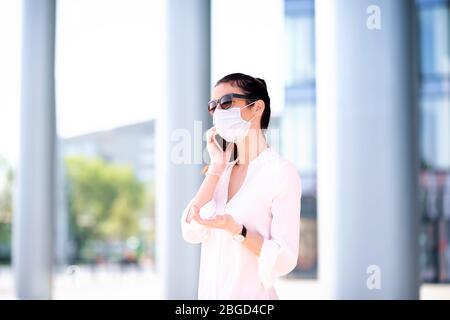 Nahaufnahme einer selbstbewussten Frau, die während coro vor dem Business Center läuft und eine Gesichtsmaske trägt und mit jemandem auf ihrem Mobiltelefon spricht Stockfoto