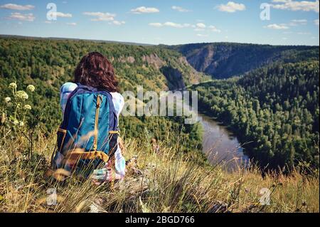 Junge glückliche Frau mit Rucksack sitzt auf einem Felsen und blickt auf das Tal darunter Stockfoto