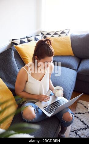 Frau, die am Laptop arbeitete, saß auf dem Sofa und trank Kaffee Stockfoto