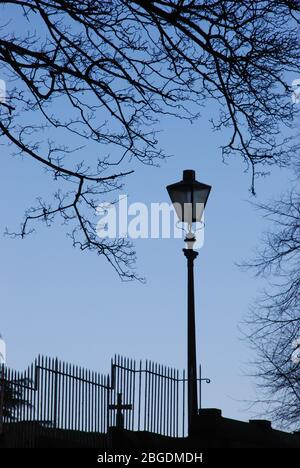 Einzelne Straßenbeleuchtung mit schmiedeeisernen versetzten Zaun und Kreuz hinter zusammen mit blattlosen Baum rund um Licht in Silhouette kurvend Stockfoto