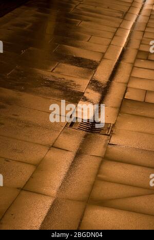 Nasse Pflasterflaggen mit zentraler Dachrinne, Abfluss und Grill alle mit einem goldenen Leuchten von reflektierten Straßenbeleuchtung Stockfoto