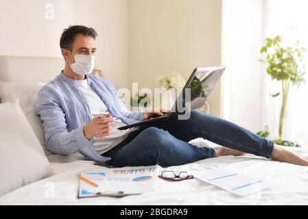 Geschäftsmann mit Gesichtsmaske in lässiger Kleidung in Hemd und Jeans, der zu Hause arbeitet, mit einem Laptop, der auf einem Bett mit Dokumenten liegt Stockfoto