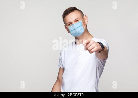 Du bist so lustig. Porträt des jungen Mannes in weißem Hemd mit chirurgischen medizinischen Maske stehend, suchen und zeigen auf Kamera mit Smiley Gesicht. Indoor st Stockfoto