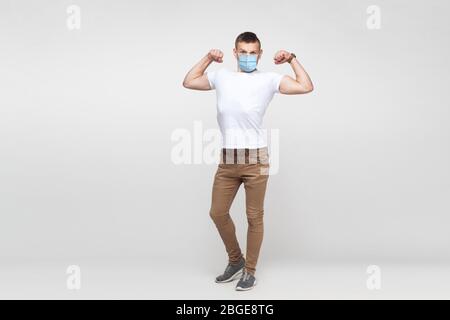 Ich bin stark. In voller Länge Porträt des jungen Mannes in weißem Hemd mit chirurgischen medizinischen Maske stehen und zeigt seine Bizeps und suchen mit hochmütigen Gesicht Stockfoto