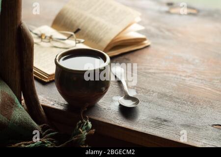 Tasse Kaffee, altes Buch und Gläser auf der Fensterbank, in der Nähe eines Stuhls mit Plaid. Retro-Style Stockfoto