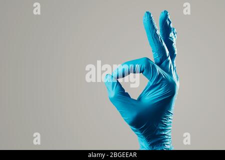 Nahaufnahme der Hand eines Mannes, der blaue Operationshandschuhe trägt, das OK-Zeichen vor einem grauen Hintergrund tut, mit etwas Leerzeichen auf der linken Seite Stockfoto