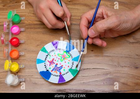 Luftaufnahme der Hände eines Erwachsenen und eines Kindes, das einen Porzellanteller mit einem Pinsel und mit einem Satz Farben in verschiedenen Farben auf Holz malt. Stockfoto