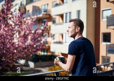 Frühlingsmorgen zu Hause. Junger Mann mit Tasse Tee oder Kaffee auf dem Balkon. Stockfoto