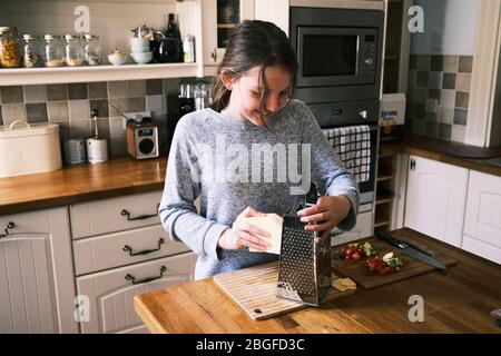 Ein junges Mädchen bereitet Essen, Reibenkäse, in einer Küche zu Hause mit Zutaten auf einer Arbeitsfläche.