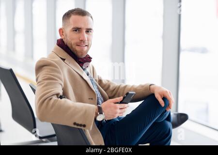 Attraktive junge Erwachsenen Mann in eleganter Schal und Sitzen im Flughafen Terminal Wartebereich Mantel. Er schaut auf zellulärer Mobiltelefon und Stockfoto