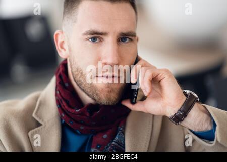 Attraktive junge Erwachsenen Mann in eleganter Schal und Sitzen im Flughafen Terminal Wartebereich Mantel. Er schaut auf zellulärer Mobiltelefon und Stockfoto
