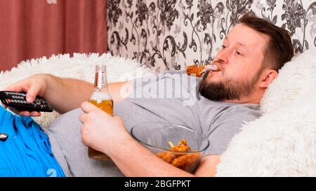Bärtiger fetter Mann mit großem Bauch liegt auf dem Sofa mit Fast Food, trinkt Bier und verpasst den Fernseher mit der Fernbedienung in der Hand, Nahaufnahme. Stockfoto