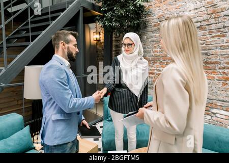 Erfolgreiche selbstbewusste muslimische Geschäftsfrau, die nach einem wichtigen Deal oder Finish mit ihren beiden kaukasischen Partnern, blonden Frauen und bärtigen Männern, die Hände schüttelt Stockfoto