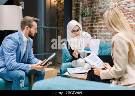 Hübsche muslimische Geschäftsfrau, Leiterin des Projekts, im Gespräch mit ihren beiden kaukasischen Kollegen, Mann und Frau, Brainstorming, Besprechung des Businessplans Stockfoto
