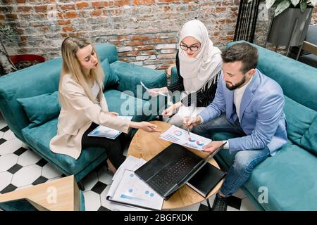Draufsicht auf die drei jungen multiethnischen Büromanager, die mit Finanzberichten und Laptop im modernen Lobbyraum arbeiten Stockfoto