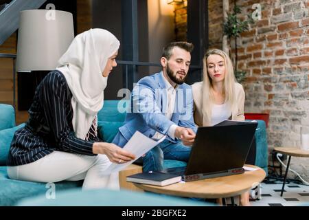 Gruppe von drei jungen gemischten Rassen Menschen, muslimische Frau, kaukasischen Mann und Frau, sitzen zusammen am Tisch mit Laptop und Projekt-Papiere Stockfoto