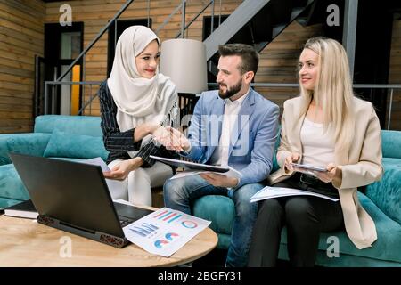 Multiethnische Gruppe von inspirierten jungen Menschen sitzen und diskutieren neues Projekt in stilvollen Loft Konferenzraum. Glücklich lächelnde muslimische Frau in Hijab Stockfoto