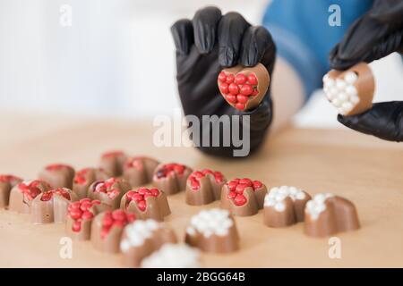 Konditor in schwarzen Handschuhen macht süße herzförmige Pralinen mit rot-weißen Füllung Stockfoto