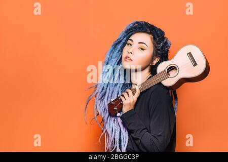 Porträt einer Hipster-Frau mit violetten Dreadlocks und Ukulele in den Händen auf orangefarbenem Hintergrund Stockfoto