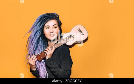 Porträt einer Hipster-Frau mit violetten Dreadlocks und Ukulele in den Händen auf orangefarbenem Hintergrund. Musikinstrument Ukulele Stockfoto