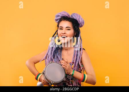 Rastafarian Frau spielt auf ethischen Mini-Trommel. Portrait Frau mit violetten Dreadlocks auf gelbem Hintergrund Stockfoto