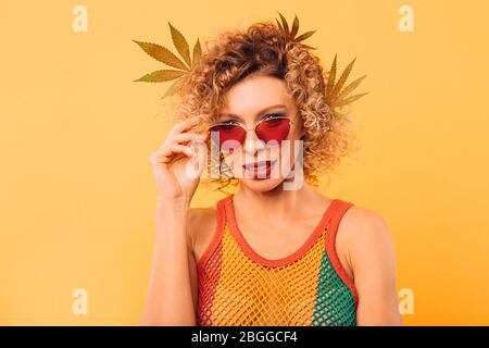Lockige Frau in rosa Brille und einem Rastakleid mit Marihuanablättern in den Haaren auf gelbem Hintergrund Stockfoto