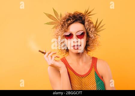 Hipster Frau raucht eine Pfeife mit Cannabis. Porträt einer Frau mit Marihuanablättern in ihren Haaren Stockfoto