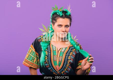 Portrait Frau mit Marihuana Blätter in ihrem Haar auf einem violetten Hintergrund. Frisur mit grünen Kanekalon. Stilvolles Hipster-Kleid Stockfoto
