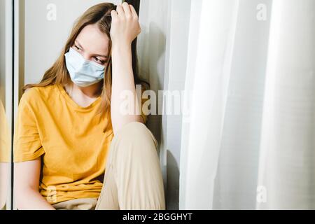 Trauriges Mädchen in medizinischer Schutzmaske und gelbem T-Shirt, das während eines pandemischen Quarantänevirus Covid-19 auf der Fensterbank sitzt. Isolation zu Hause Stockfoto