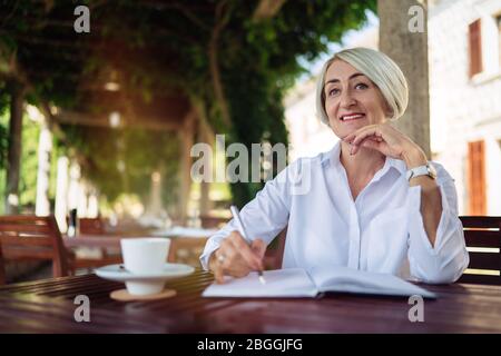 Glückliche ältere Frau, die in ein Notizbuch oder Tagebuch in einem Café schreibt Stockfoto