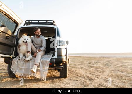 Schöner junger bärtiger Mann sitzt im hinteren Teil seines Autos und spielt mit Hund am Strand Stockfoto