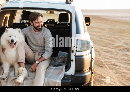 Schöner junger bärtiger Mann sitzt im hinteren Teil seines Autos und spielt mit Hund am Strand Stockfoto