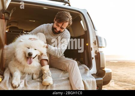 Schöner junger bärtiger Mann sitzt im hinteren Teil seines Autos und spielt mit Hund am Strand Stockfoto