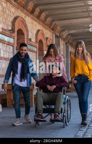 Älterer Mann mit Brille und paralytischem Hut im Rollstuhl, begleitet von einer jungen lateinamerikanischen und zwei kaukasischen Mädchen, die glücklich die Stre hinunterrollen Stockfoto