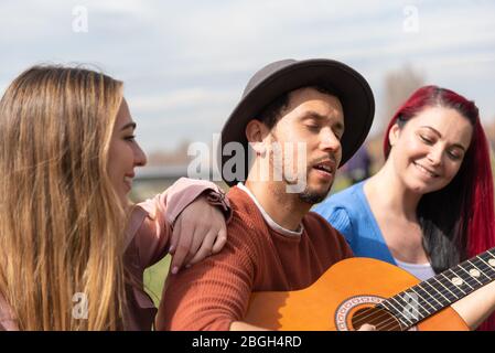 Ein hispanischer Junge in einem Hut spielt neben zwei kaukasischen Mädchen in einem Stadtpark die Gitarre Stockfoto