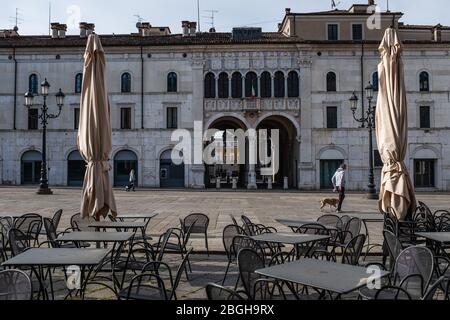 Zwei Leute gingen über den Hauptplatz in Brescia (Italien). Menschenleere Tische vor einem Restaurant während der Coronavirus-Notabsperrung. Stockfoto