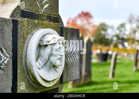 Friedhof im Herbst. Detail eines alten grauen Grabsteins auf einem Friedhof im Herbst mit dem Gesicht von Jesus Christus in weißem Marmor gegen einen blauen Himmel. Stockfoto