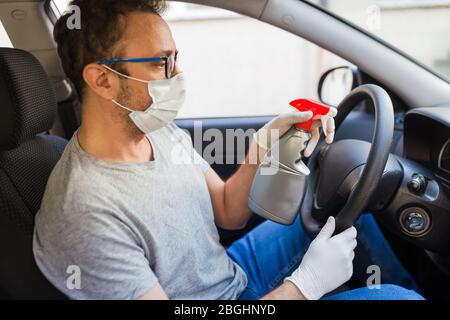 Fahrer trägt chirurgische Maske und Schutzhandschuhe beim Sprühen Desinfektionsmittel auf Auto Lenkrad zu Infektionen zu verhindern. Stockfoto