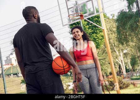 Outdoor-Aktivitäten. Afrikanisches Paar stehend auf Basketballplatz Mann Rückansicht mit Ball auf Frau zu Fuß in Richtung Lachen spielerisch Stockfoto
