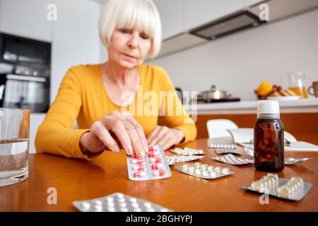 Ruhestand. Ältere Frau sitzt am Tisch in der Küche unter Blister mit Pillen Gefühl unwohl Nahaufnahme besorgt Stockfoto