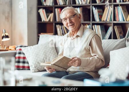 Senior Mann allein sitzt in der Bibliothek in Brillen Buch halten Blick Kamera neugierig Stockfoto