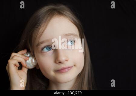 Nettes lächelndes blondes Mädchen, das Muschelschale neben ihrem Ohr hält und dem Ozean auf schwarzem Hintergrund im Studio lauscht Stockfoto