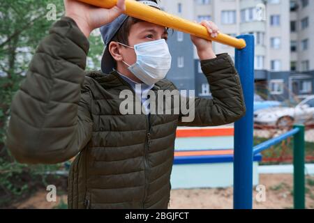 Teenager-Junge steht in der Nähe horizontale Bar von Outdoor-Trainingsgeräten, auf Spielplatz in der Nähe von Hochhäusern mit Wohnungen, eine medizinische Maske auf seinem Gesicht p Stockfoto