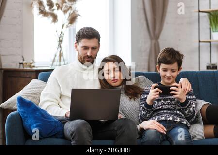 Mutter, Vater und Sohn mit Laptop und Handy zu Hause. Familienmitglieder ignorieren einander und sprechen live. Gadget Einfluss auf Familie Stockfoto