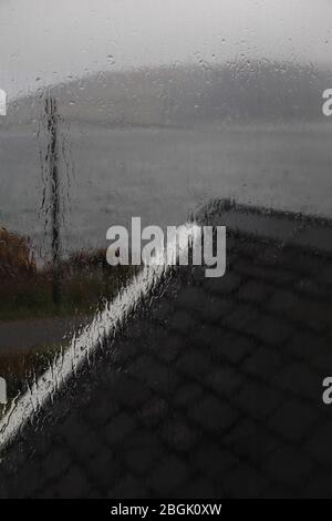 Blick aus einem regnerischen Fenster, das Wasser läuft hinunter, über ein schwarzes Schieferdach mit weißen Zierleisten zu einem loch + entfernten Hügel an einem dunklen, trostlos schottischen Tag Stockfoto